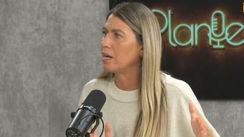 Una mujer de cabello rubio habla frente a un micrófono negro en un estudio con un letrero luminoso al fondo