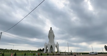 Estatua blanca de figura humana en una base con arcos, bajo un cielo nublado. Se observan cables eléctricos, postes, árboles, pasto, personas y una furgoneta blanca