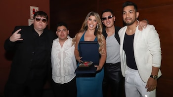 Yahaira Plasencia, sonriente y con un vestido azul brillante, sostiene un premio dentro de una caja azul, rodeada por cuatro hombres en un evento
