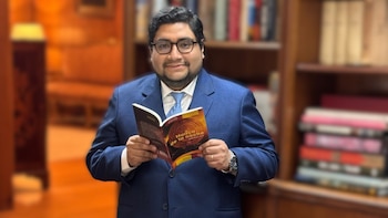 Hombre sonriente con traje azul y gafas sostiene un libro abierto con la portada naranja y el título "Huaca de la noche". Detrás hay una estantería