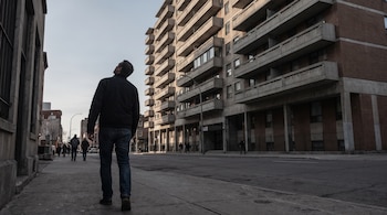 Un hombre visto de espaldas camina por una vereda de concreto frente a un edificio alto de ladrillo y hormigón con balcones.