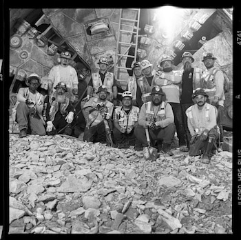 Fotografía en blanco y negro de trece trabajadores, con cascos y chalecos reflectantes, posando frente a la cabeza de una tuneladora, rodeados de escombros de roca
