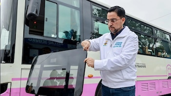 Un hombre con chaqueta blanca sostiene un panel de vidrio oscuro frente a un autobús blanco con una franja rosada