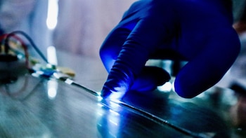 Una mano con un guante azul presiona una delgada tira sobre una superficie, donde se enciende una luz brillante bajo el dedo