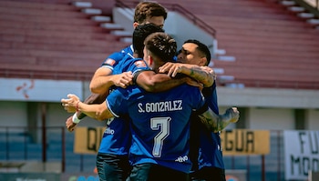 Cuatro futbolistas con camiseta azul se abrazan en un estadio de fútbol