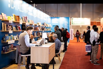 Personas conversan y trabajan en un stand de libros azul con estanterías llenas, mientras otras caminan por una alfombra roja en un evento literario