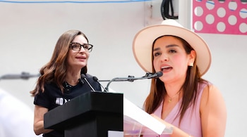 Dos mujeres en primer plano; una con gafas y camiseta negra, la otra con sombrero, blusa rosa, cadena y pendientes; ambas frente a micrófonos y atril.