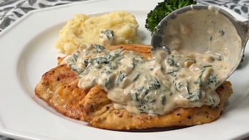 Plato blanco con suprema de pollo, puré de patatas y brócoli. Una cuchara metálica vierte salsa cremosa de espinaca. Fondo con mantelería de hojas, casas decorativas y un árbol pequeño.