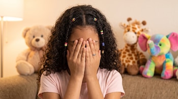 Una niña de cabello rizado cubre su rostro con las manos en un sofá junto a un oso, una jirafa y un elefante de peluche.