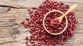 Un conjunto de frijoles rojos esparcidos sobre una mesa de madera, con un cuenco de madera lleno de frijoles rojos y una cuchara de madera también con frijoles.
