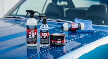 Cuatro productos de limpieza automotriz, incluyendo jabón, detallador rápido y cera, junto a una toalla de microfibra azul, sobre el capó brillante de un coche azul.