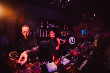 En el interior de un bar con luz roja, un hombre y una mujer preparan cócteles en la barra, donde se ven botellas y varias copas con bebidas rosadas