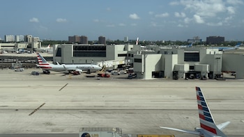 Avión blanco de American Airlines siendo empujado por un tractor azul en la pista de un aeropuerto soleado, con un trabajador y edificios en el fondo
