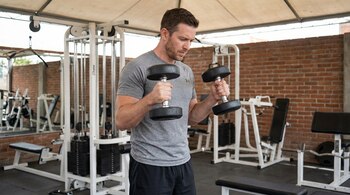 Hombre de unos 30 años, vestido con camiseta gris y pantalón corto negro, de pie en un gimnasio, realizando curl de bíceps martillo con dos mancuernas.