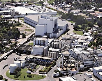 Vista aérea de una gran planta industrial blanca con múltiples edificios, tanques cilíndricos y camiones, rodeada de vegetación y zonas urbanas
