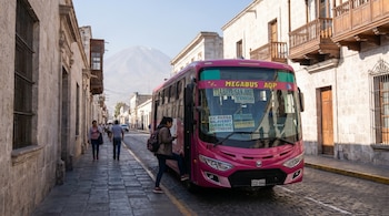 Mujer subiendo a un bus urbano rosa en una calle empedrada de Arequipa, Perú. Edificios de sillar blanco y el volcán Misti son visibles al fondo.
