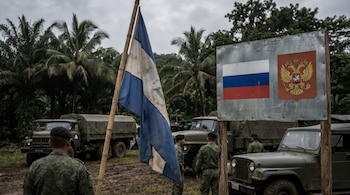 Imagen de un puesto militar en el trópico. Soldados uniformados, vehículos militares, bandera de Nicaragua y cartel con bandera y escudo de Rusia bajo cielo gris.