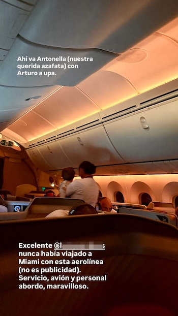 Interior de un avión de pasajeros con luz cálida. Una azafata vestida de blanco sostiene a un niño pequeño. Se ven asientos de primera clase y pasajeros