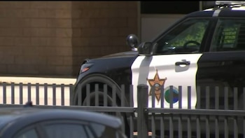 Vista lateral de um carro de polícia SUV preto e branco com marcações de xerife, parcialmente escondido por uma cerca escura, com um prédio de tijolos ao fundo.