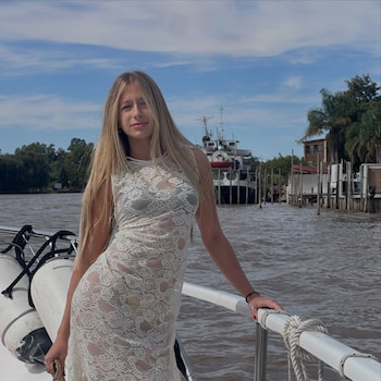Una joven con cabello largo rubio y un vestido de encaje blanco en la cubierta de un barco con un río, otras embarcaciones y árboles al fondo