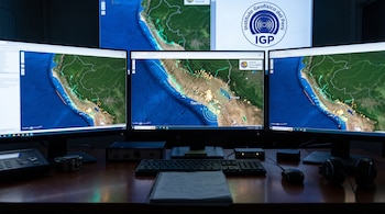 Varios monitores con mapas sísmicos sobre un escritorio con teclado, libreta de notas, ratón y auriculares en una sala técnica.