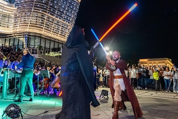 Dos personas disfrazadas, una de Darth Maul y otra de Obi-Wan Kenobi, luchan con sables de luz. Detrás, una orquesta y un edificio moderno iluminado