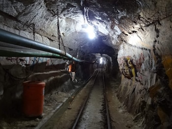 Interior de un túnel de roca con vías de ferrocarril, dos tuberías suspendidas, cadenas, malla metálica, barril naranja y luces en el techo