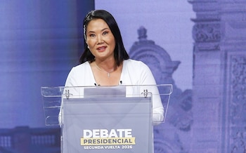 Keiko Fujimori, una mujer de cabello oscuro, habla en un podio transparente con el texto "DEBATE PRESIDENCIAL SEGUNDA VUELTA 2026". Viste una blusa blanca y un collar con una cruz plateada. El fondo es púrpura y borroso con detalles arquitectónicos