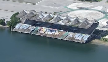 Vista aérea de un estadio deteriorado junto a una extensión de agua, con gradas y techo dañados. Hay una gran explanada pavimentada alrededor