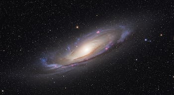 Imagen de una galaxia espiral vista desde el espacio, con un núcleo brillante y brazos tenues de estrellas, polvo y gas, sobre un fondo de innumerables estrellas.