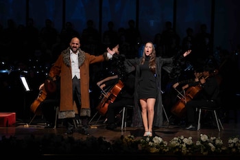 Un hombre con abrigo marrón y una mujer con vestido negro cantan sobre un escenario frente a una orquesta