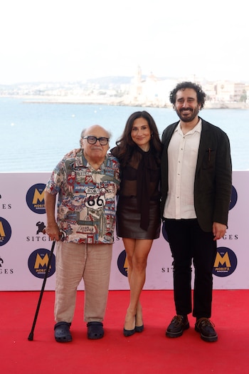 Danny DeVito con bastón, Lucy DeVito y Jake DeVito posan sobre una alfombra roja ante un panel publicitario con el mar al fondo