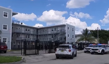Un edificio de tres pisos pintado de gris con rejas en el frente y varios vehículos de la policía estacionados en la calle