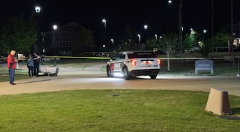 Vehículo policial y cinta amarilla de precaución en el campus de Arkansas State University de noche; reporteros y cámaras presentes bajo farolas brillantes