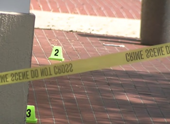 Cinta amarilla con texto 'CRIME SCENE DO NOT CROSS', marcadores de evidencia verdes con los números dos y tres, y suelo de ladrillos rojos