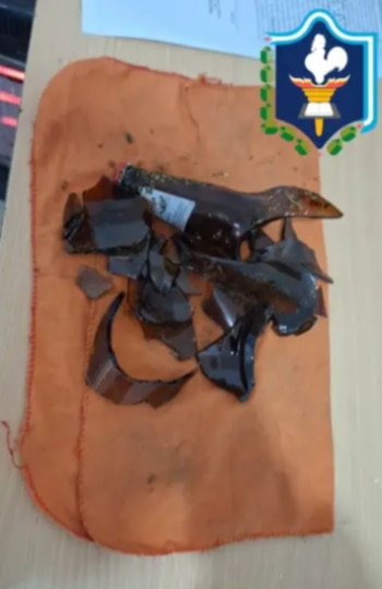Fragmentos de una botella de vidrio oscura rota sobre un paño de tela naranja. Un escudo con un gallo blanco y una antorcha aparece en la esquina superior derecha