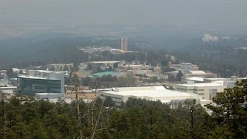 Vista aérea de un complejo de edificios del Laboratorio Nacional Los Álamos entre densos bosques de coníferas bajo un cielo nublado y brumoso