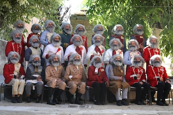 Diecinueve hombres con pelucas rizadas grises y barbas grises, vestidos con trajes de chef, uniformes militares y otras prendas, posan ante vegetación