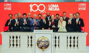 Grupo de personas aplaudiendo en el podio de la Bolsa de Nueva York, con un mazo y una campana dorada. Pantalla roja de fondo con el logo de Loma Negra y el texto 100 Años