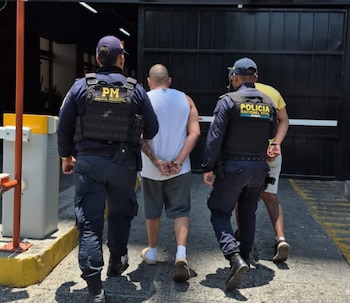 Vista trasera de dos hombres esposados, uno en camiseta blanca, otro en camisa amarilla, siendo escoltados por dos agentes de policía en uniformes oscuros