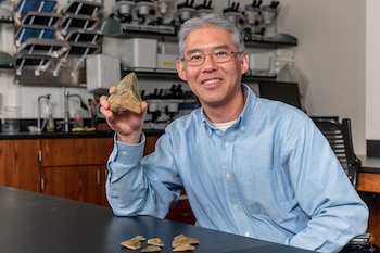 Un hombre con gafas y camisa azul sostiene un diente fósil grande. Sobre una mesa negra hay dientes fósiles pequeños y al fondo se ve un laboratorio