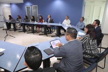 Un grupo de diez profesionales sentados alrededor de una mesa de conferencias en forma de U, con laptops y botellas de agua. Las paredes son azules