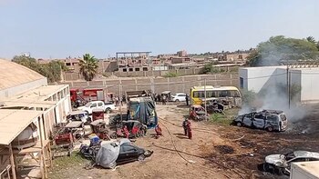 Vista panorámica de un depósito con vehículos calcinados, humo, personal de bomberos, un camión de bomberos, un bus amarillo y edificios