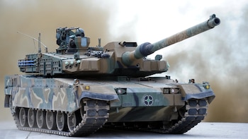 Tanque militar K2 Black Panther de camuflaje verde, marrón y negro con cañón largo, torreta, ametralladora y orugas, sobre un fondo de humo o polvo