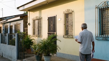 Hombre de espaldas con documentos frente a casa de fachada amarilla y azul, con rejas en ventanas y puerta, macetas con plantas y un muro.