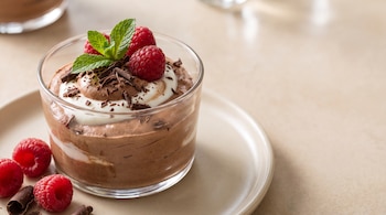 Vaso de cristal con mousse de chocolate y crema blanca, decorado con virutas de chocolate, hojas de menta y frambuesas rojas sobre un plato beige.