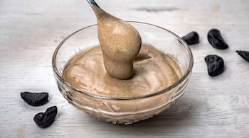 Un cuenco de vidrio con salsa cremosa, una cuchara de la que cae aderezo y varios dientes de ajo negro sobre una mesa de madera blanca.