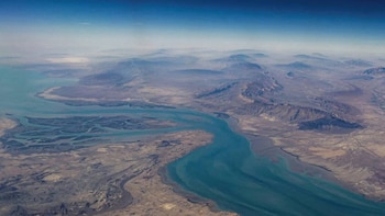 Vista aérea de un estrecho azul turquesa serpenteando entre tierra árida marrón y montañas rocosas, con el cielo azul claro difuminándose en la bruma del horizonte