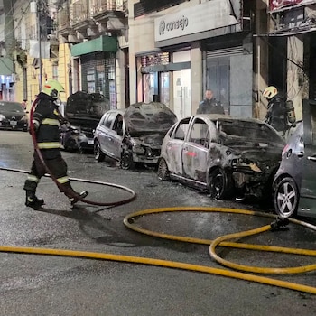 Dos bomberos, un oficial de policía, varios vehículos subcompactos y hatchback quemados, y mangueras amarillas sobre asfalto en una calle de noche