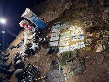 Vista aérea de varias placas de vehículos, equipo táctico, bolsas con paquetes y dos esferas oscuras en el suelo de tierra, iluminado por una linterna